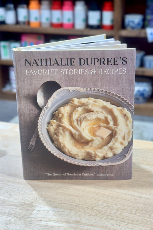 Book titled 'Nathalie Dupree's Favorite Stories & Recipes' on a wooden surface with a blurred background of shelves.