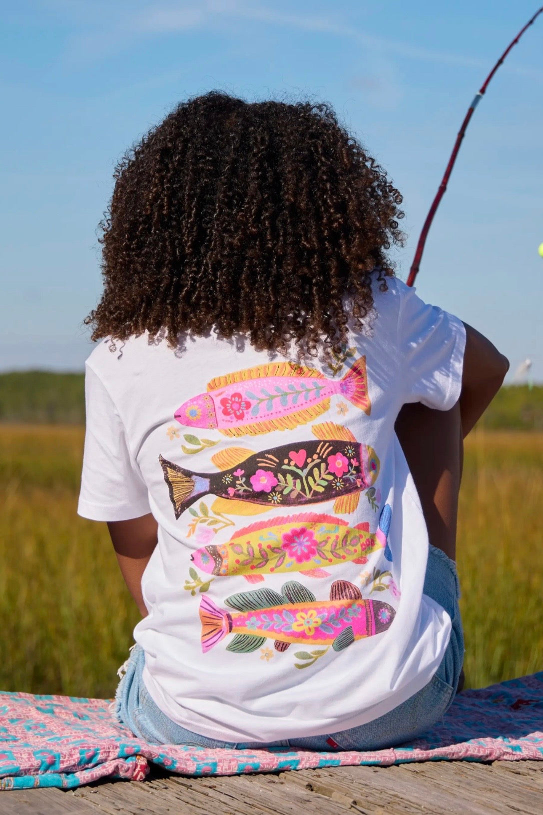 Person wearing a white t-shirt with colorful fish designs sitting on a wooden dock.