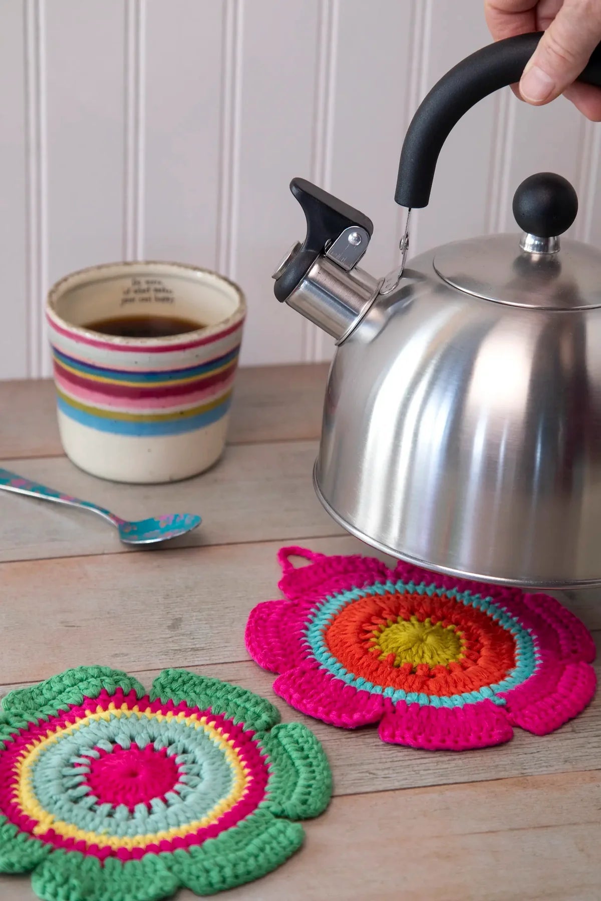 Colorful crochet trivets on a wooden surface with a teakettle and coffee cup.