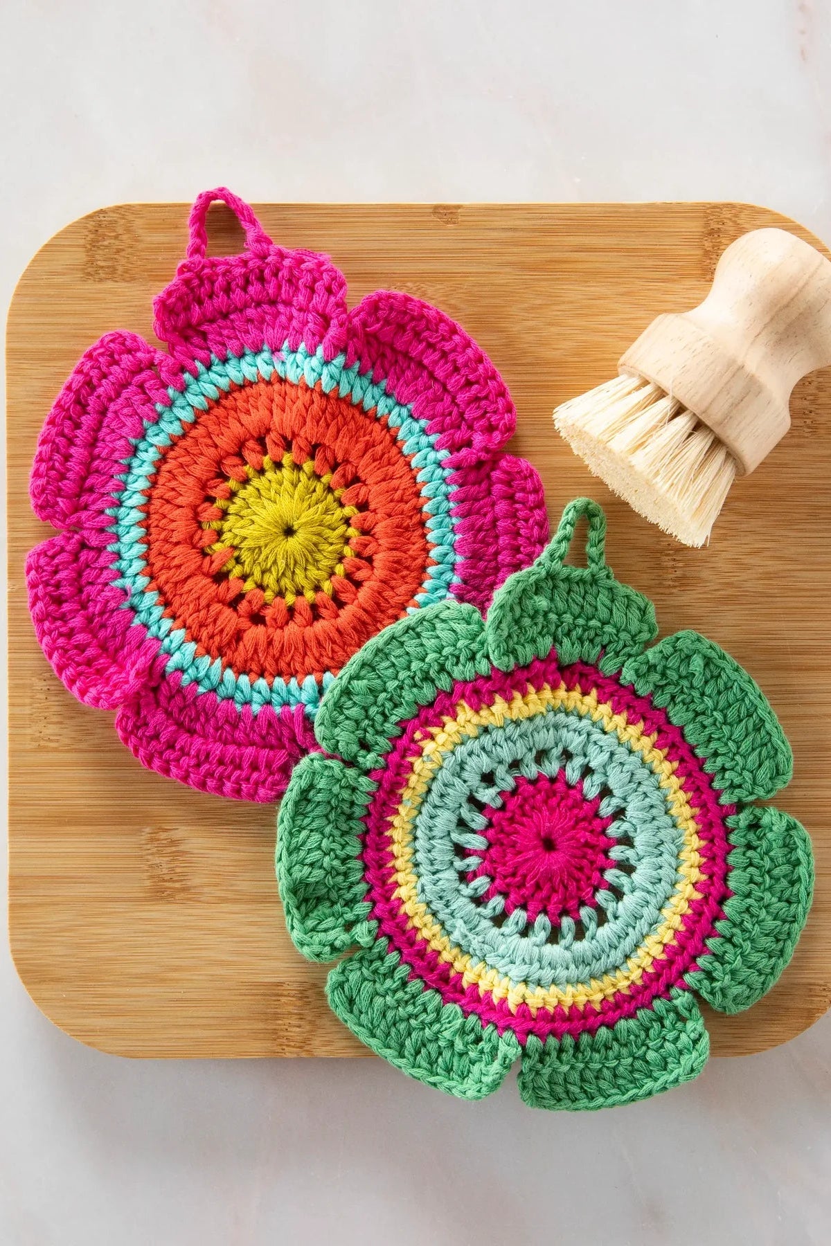 Two colorful crocheted pot holders on a wooden board with a scrub brush. Natural Life Daisy Crochet Dishcloths/Trivets, Set of 2 .