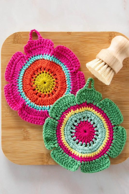 Two colorful crocheted pot holders on a wooden board with a scrub brush. Natural Life Daisy Crochet Dishcloths/Trivets, Set of 2 .