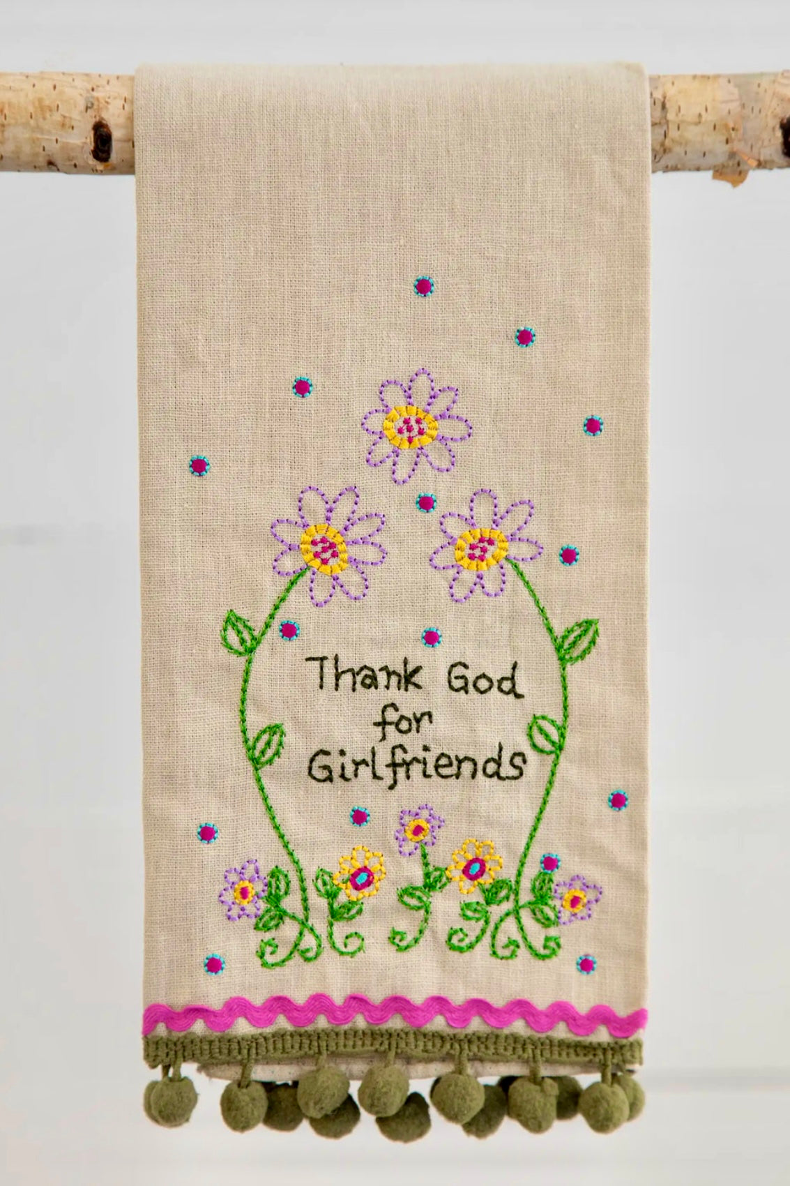 A beige linen hand towel with floral embroidery and the text 'Thank God for Girlfriends' displayed. It also features green pom-pom trim at the bottom edge.
