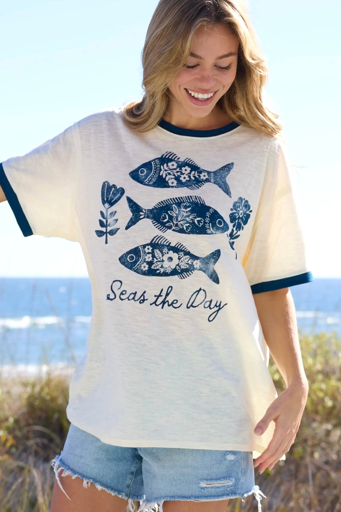 Woman wearing a t-shirt with fish design and 'Seas the Day' text by the beach.