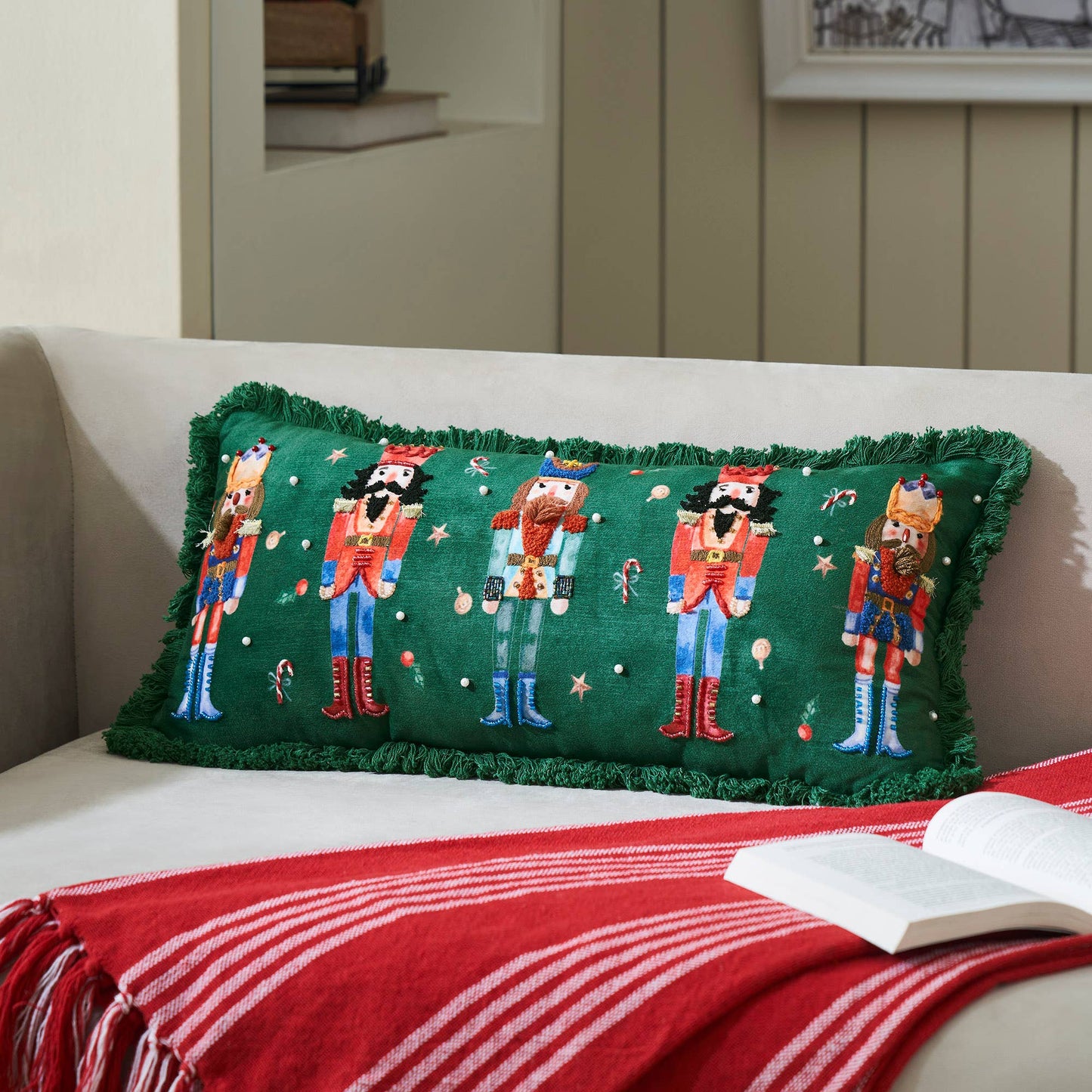 Decorative pillow with nutcracker design on a couch with a red blanket and open book.