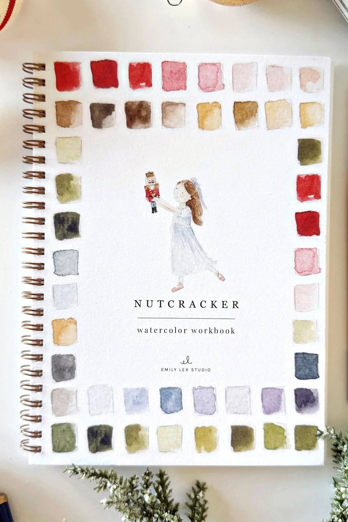 Watercolor workbook titled 'Nutcracker' with a pen and Christmas decorations on a white surface