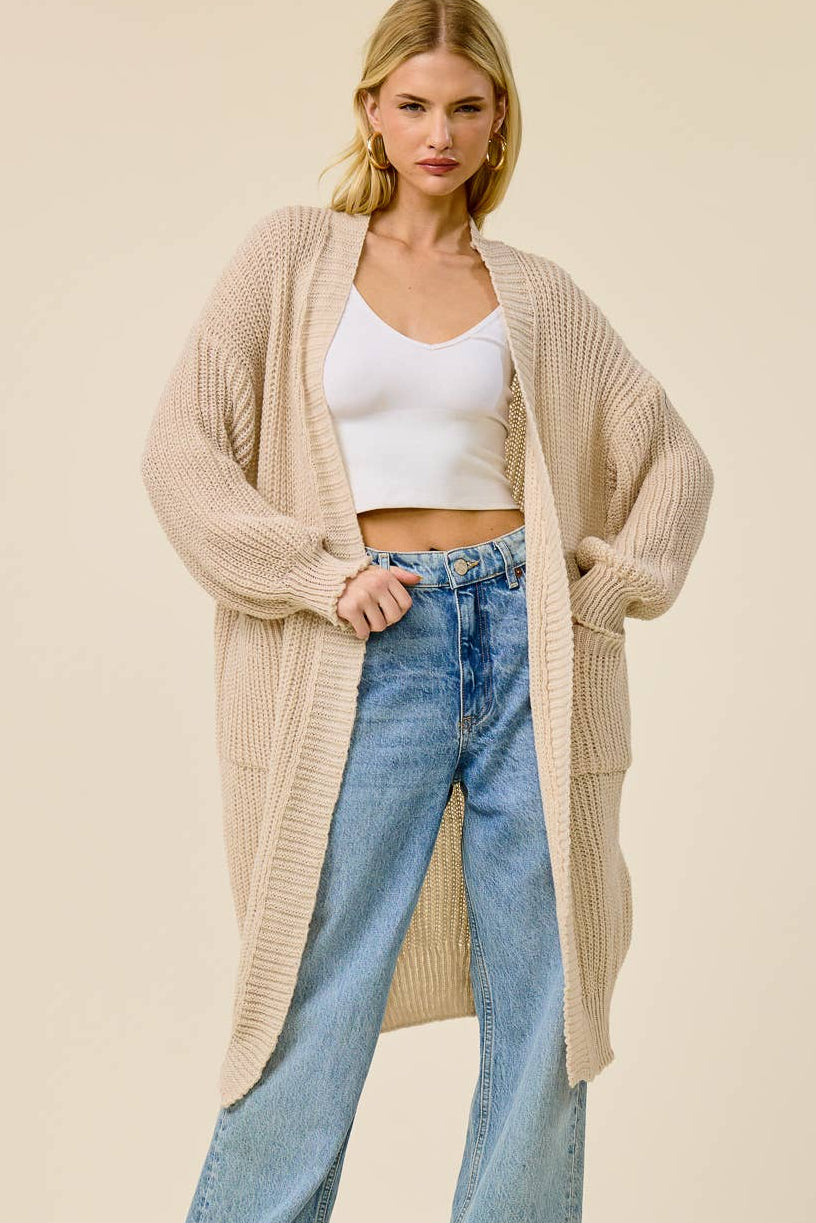 A woman modeling a long, open-front ribbed knit cardigan in a light beige color, worn over a white crop top and light-wash blue wide-leg jeans, against a solid beige studio background.