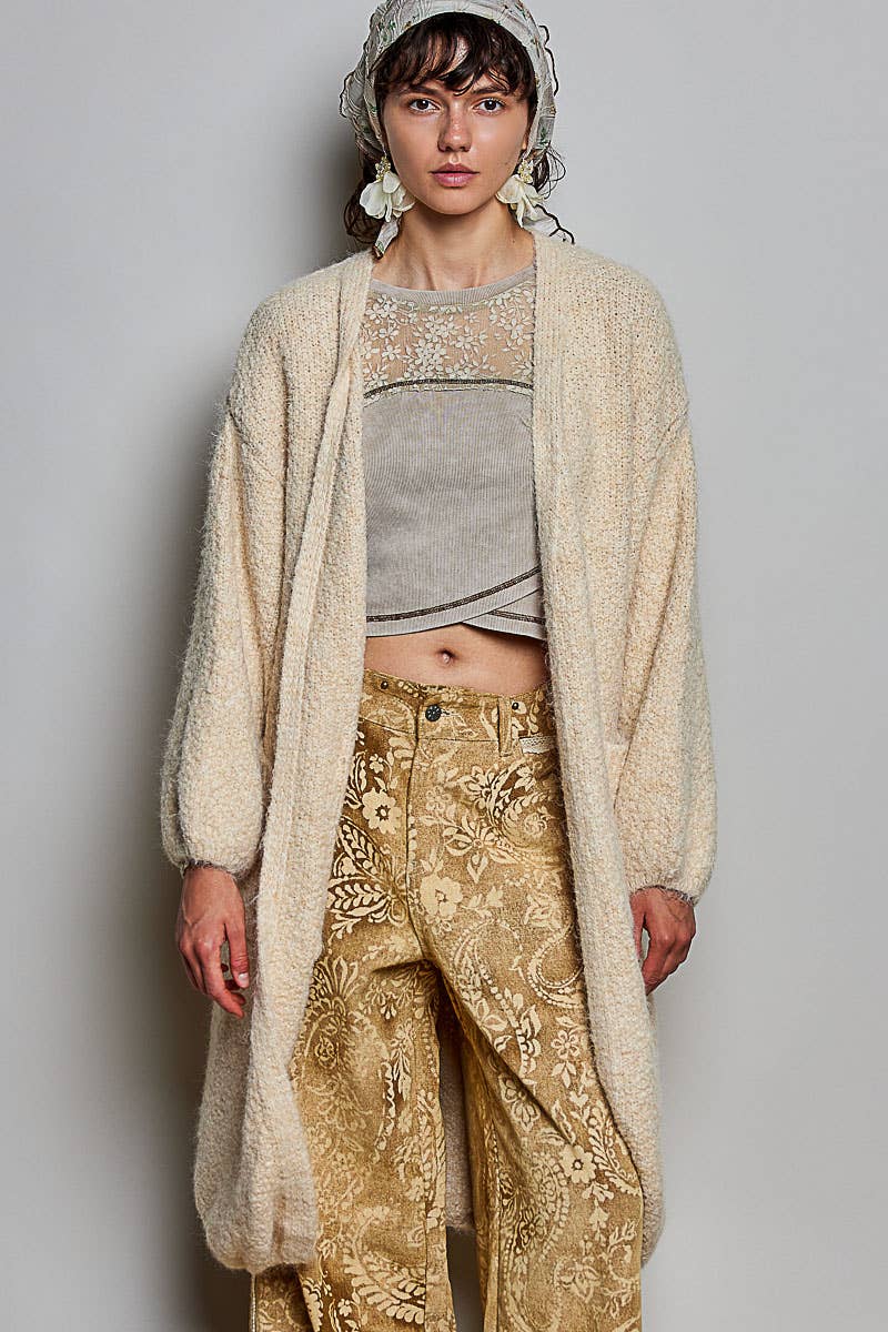 A model wearing a cream-colored, long-sleeved, open-front fuzzy cardigan over a gray cropped top and gold paisley trousers, styled with a matching headscarf.