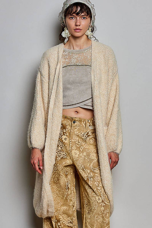A model wearing a cream-colored, long-sleeved, open-front fuzzy cardigan over a gray cropped top and gold paisley trousers, styled with a matching headscarf.