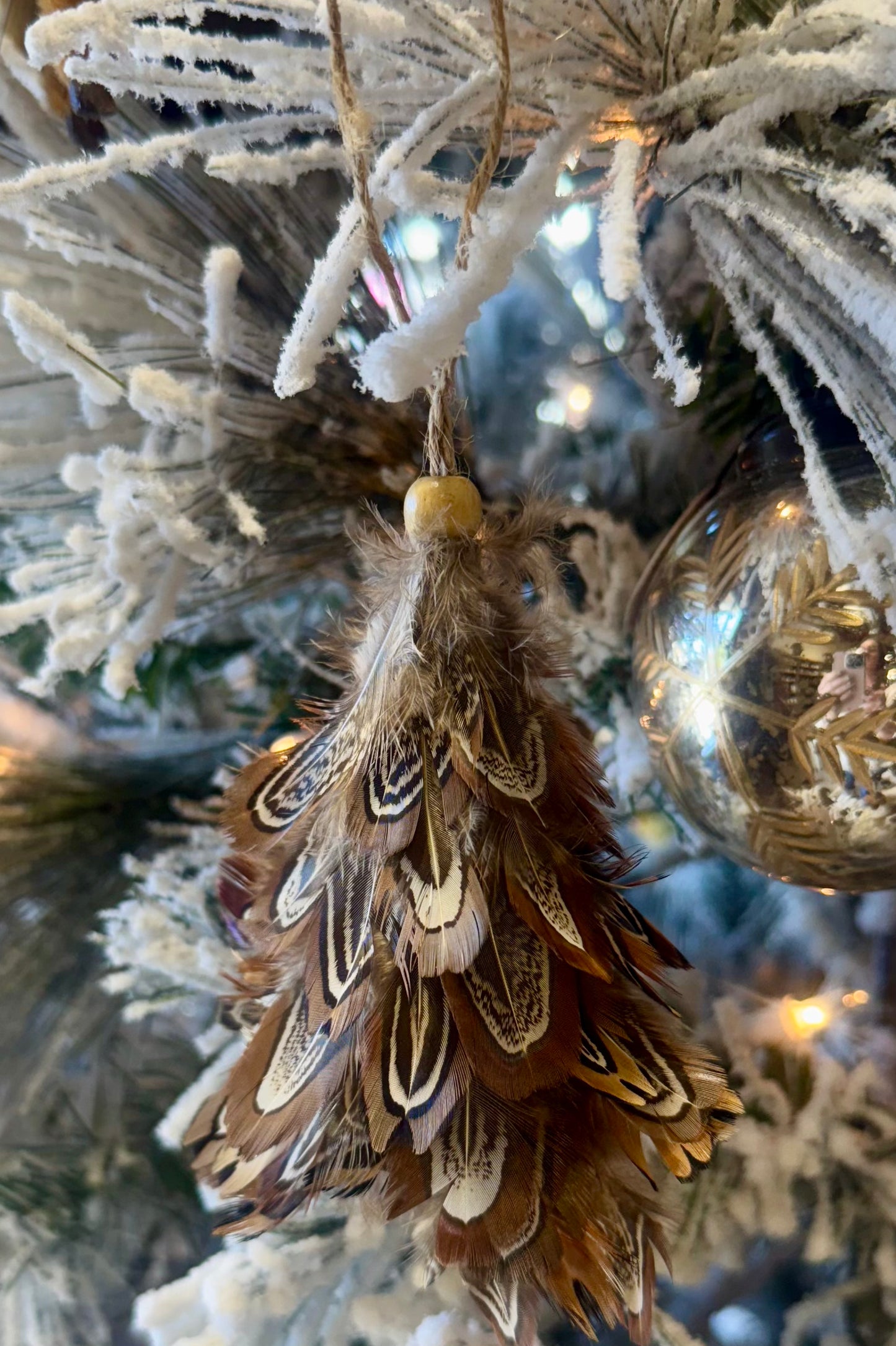 Decorative feather ornament hanging on a frosted Christmas tree with blurred lights in the background.