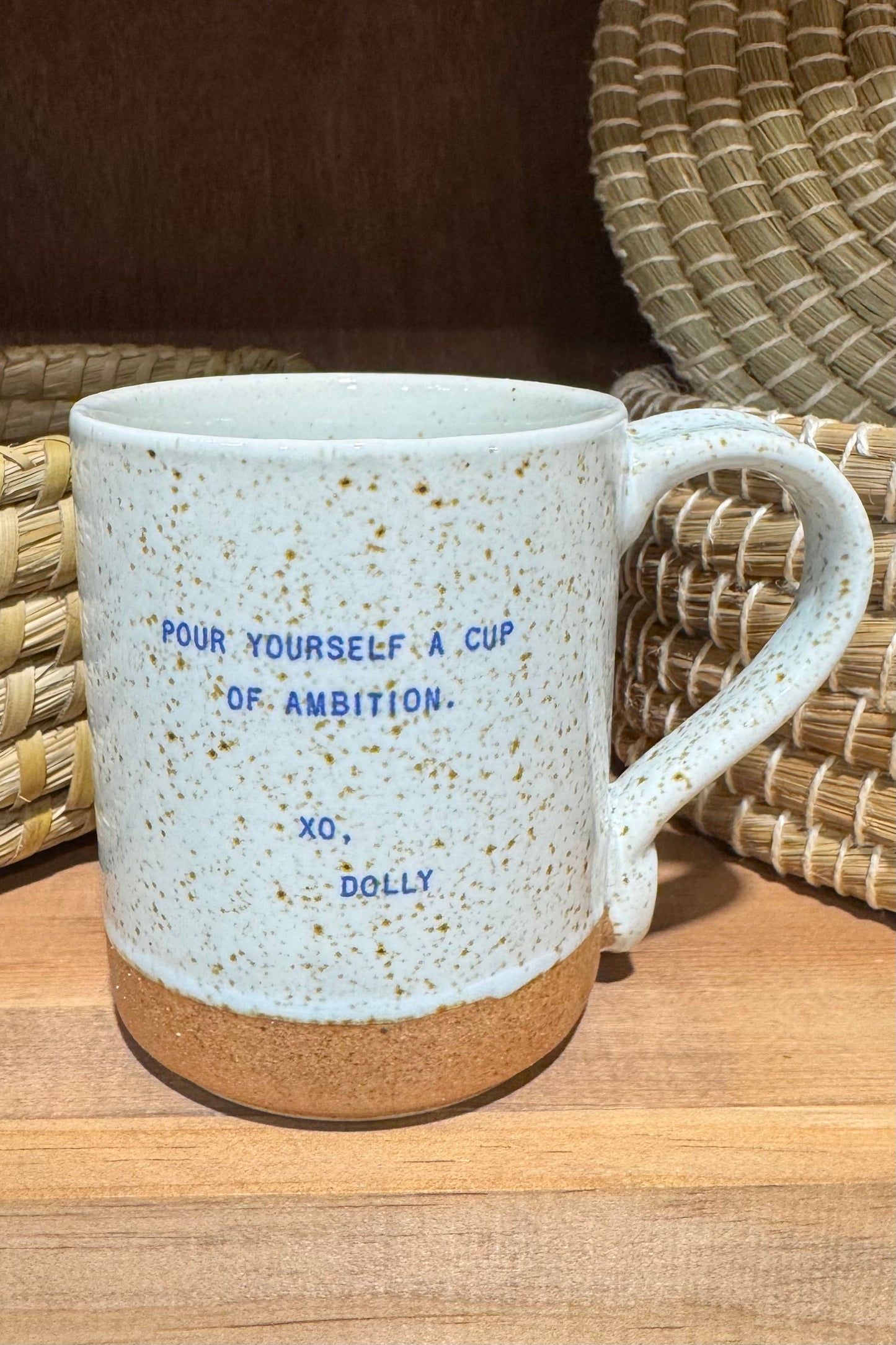 A speckled ceramic mug, featuring the text 'POUR YOURSELF A CUP OF AMBITION. XO, DOLLY' in blue.