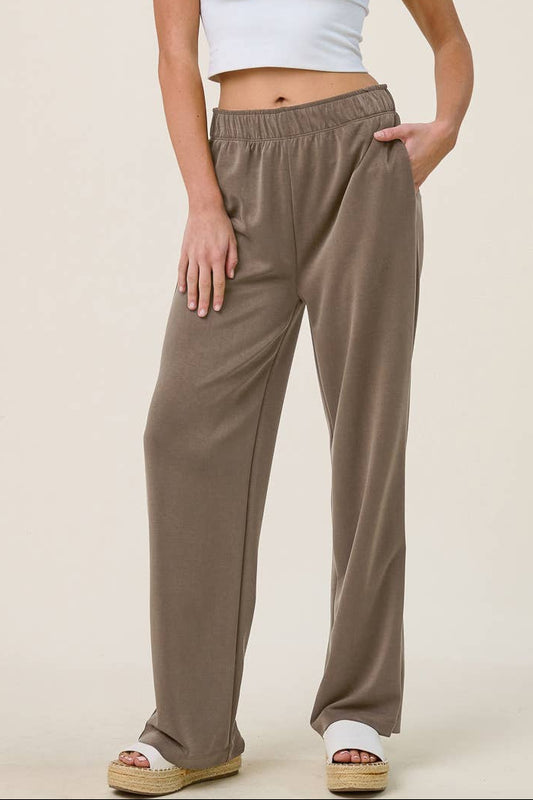 A woman modeling a white ribbed crop tank top and loose taupe-brown wide-leg sweatpants against a solid light beige studio background.