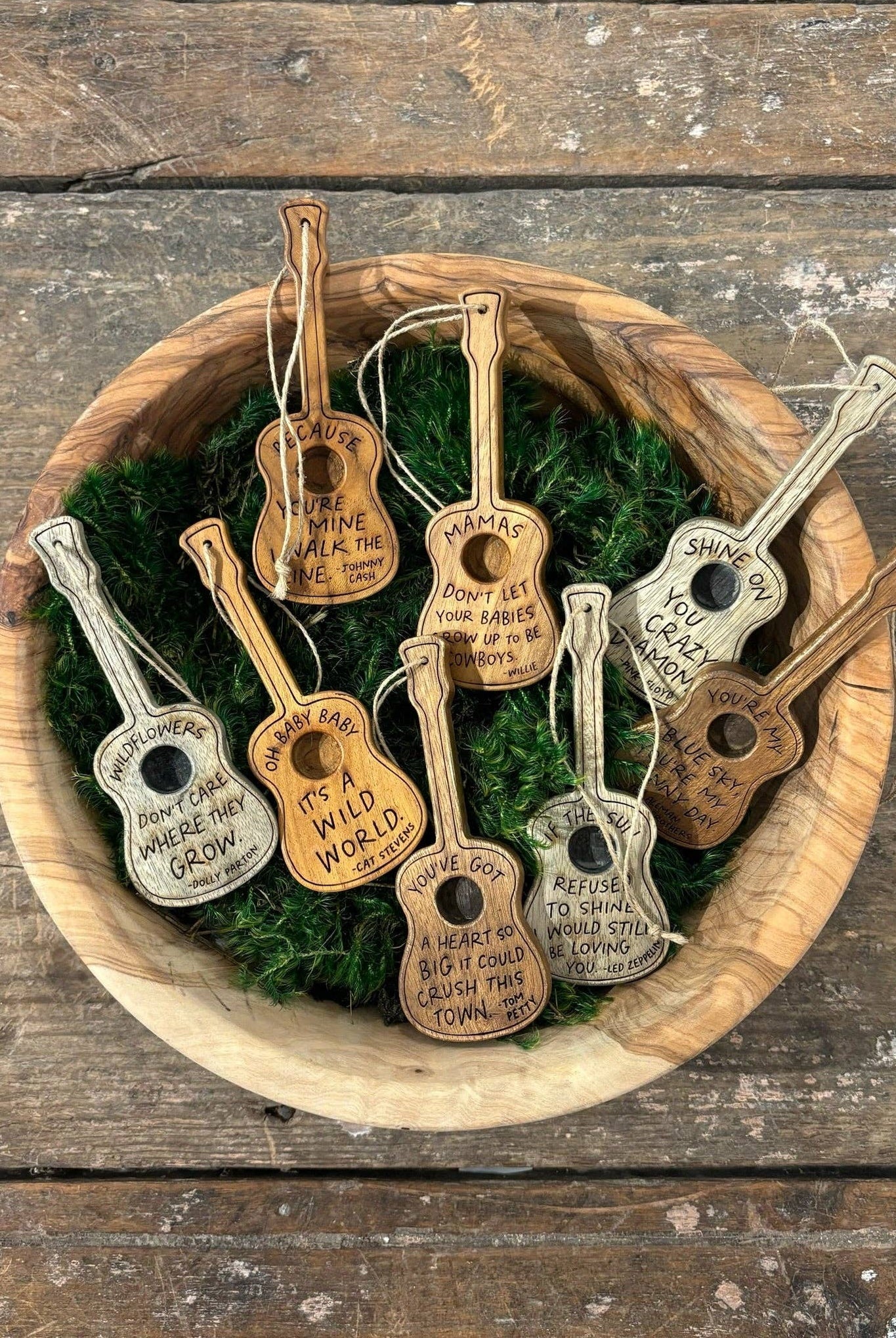 A collection of miniature guitar-shaped ornaments with engraved inspirational quotes, displayed in a wooden bowl filled with green moss on a rustic wooden tabletop.