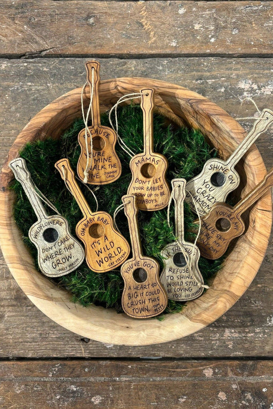 A collection of miniature guitar-shaped ornaments with engraved inspirational quotes, displayed in a wooden bowl filled with green moss on a rustic wooden tabletop.