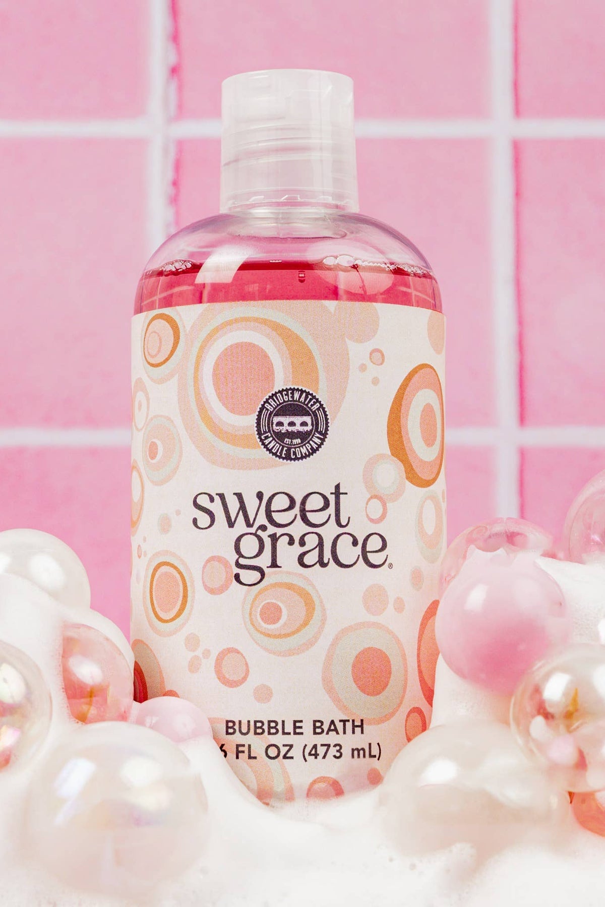 A bottle of Sweet Grace Bubble Bath with pink liquid, surrounded by white and iridescent bath bubbles against a pink tiled background.