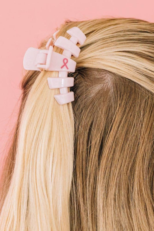 Person with blonde hair wearing a pink hair clip with a breast cancer awareness ribbon on a pink background