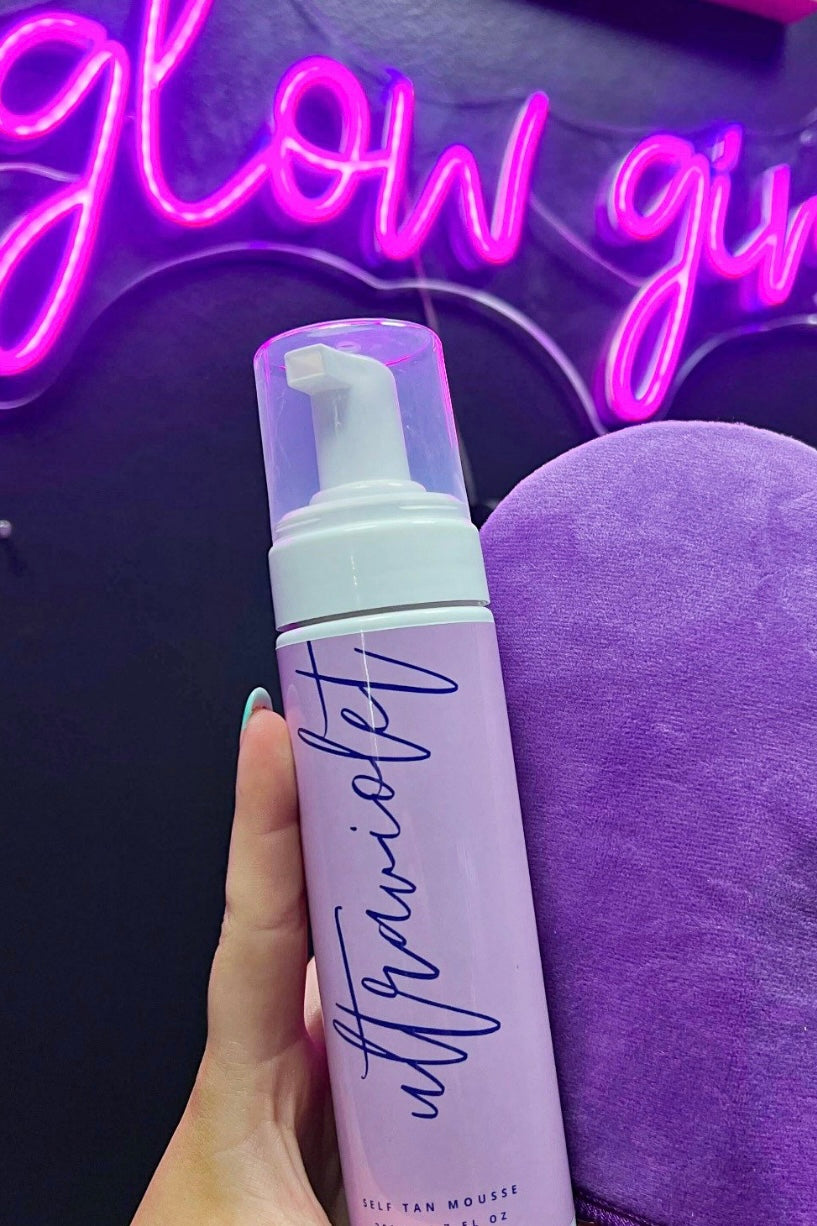 A hand holding a pump bottle of Ultraviolet Premium Self Tanning Foam, with a purple mitt and neon pink sign in the background.
