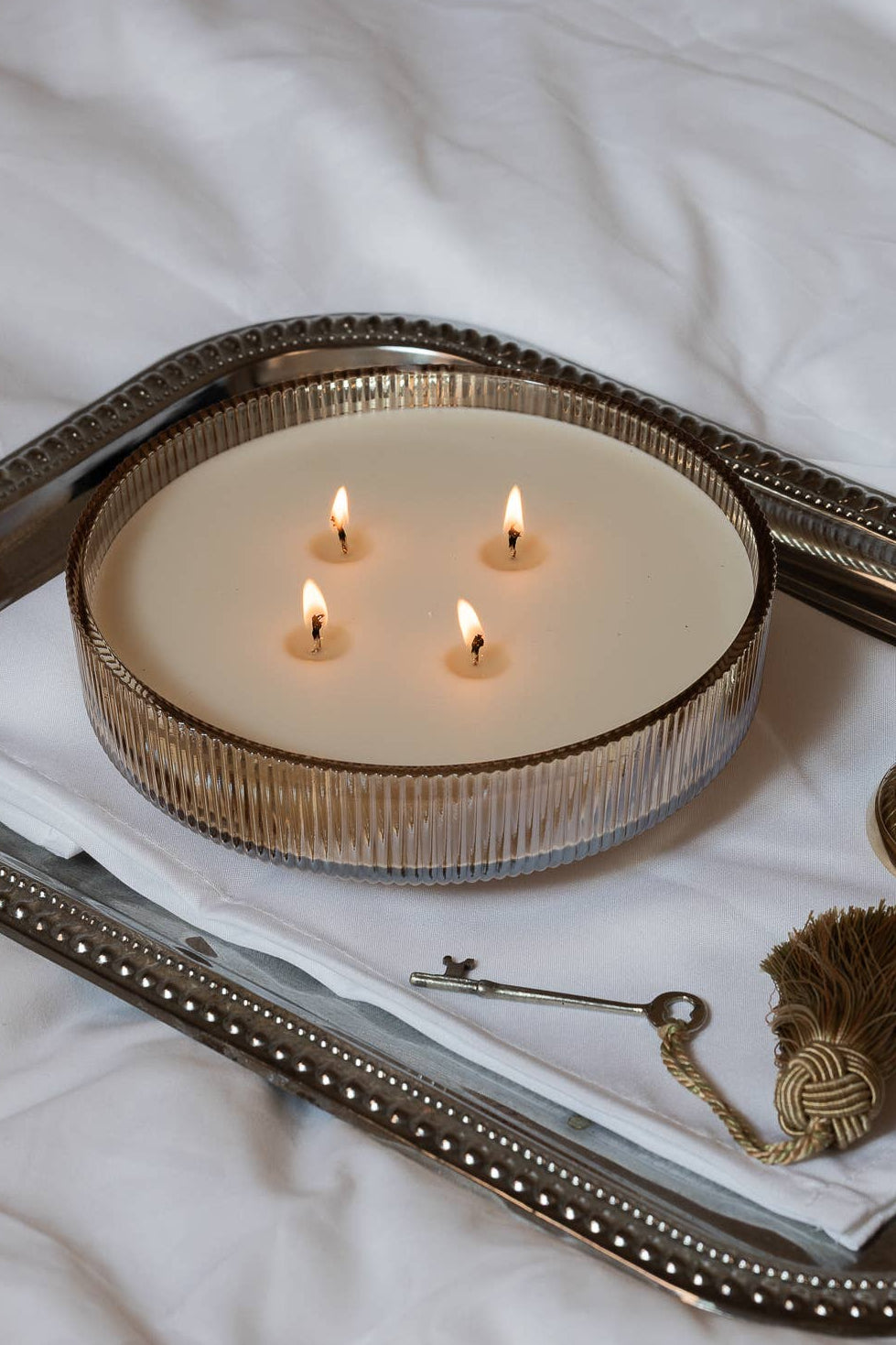 A decorative silver tray with a beaded edge holds a lit cream candle in a ribbed glass holder, alongside a small gold-toned bell, a vintage key, and a feather ornament, all arranged on white fabric. Velvet Rouge Holiday Multi-Wick Candle- 26.4 oz