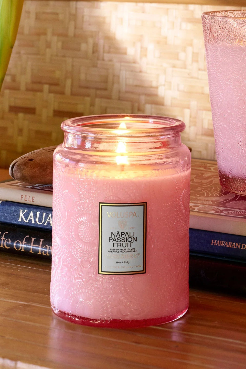 Pink candles with labels on a wooden surface with books and a mirror in the background. Voluspa Napali Passion Fruit 18 0z Large Jar Candle