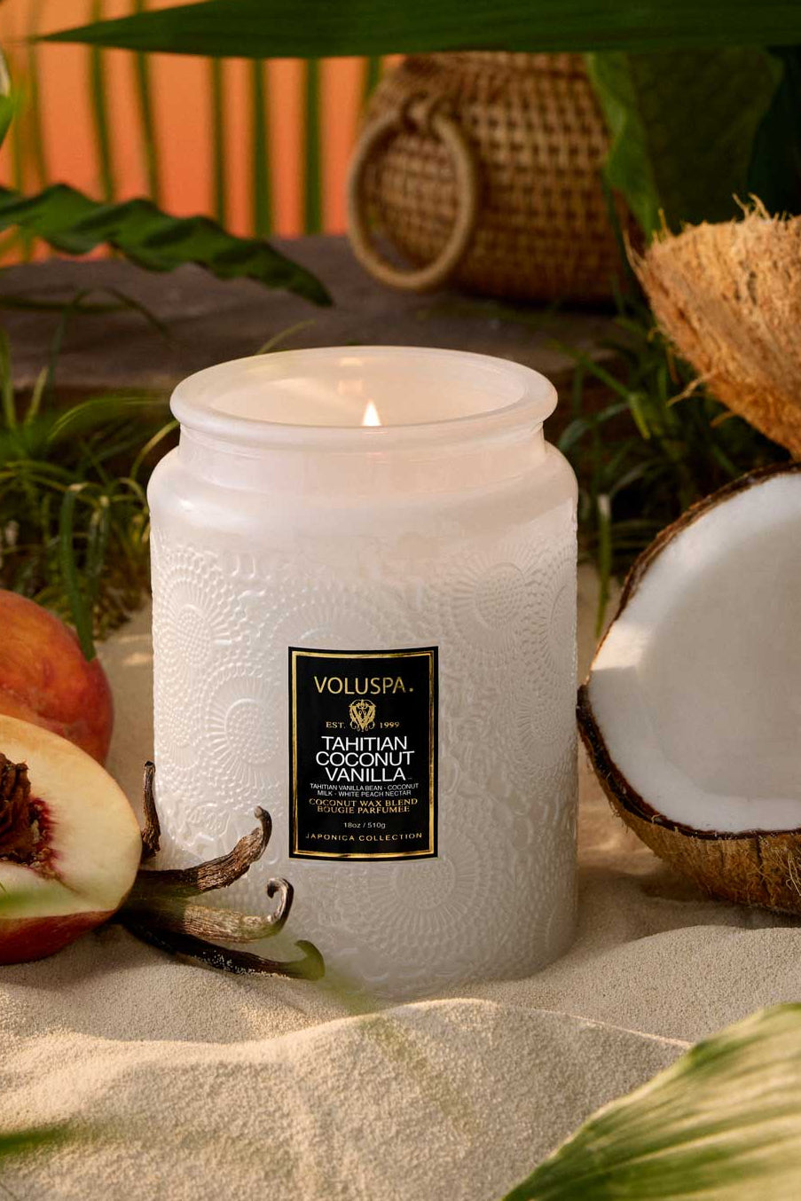 White candle with a label on a sandy surface surrounded by peaches and coconuts, with a blurred natural background. Voluspa Tahitian Coconut 18 0z Large Jar Candle