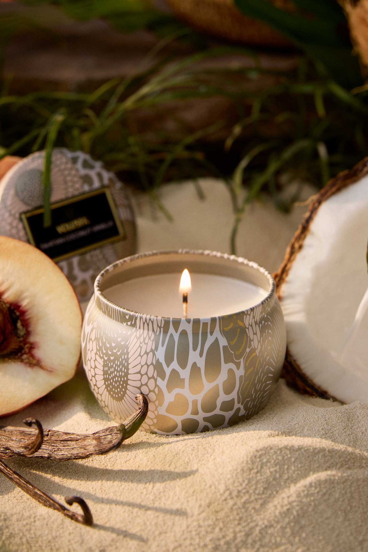 Candle in a decorative holder with peaches, a coconut, and a vanilla bean on a sandy surface. Voluspa Tahitian Coconut Vanilla 4 oz Mini Tin Candle