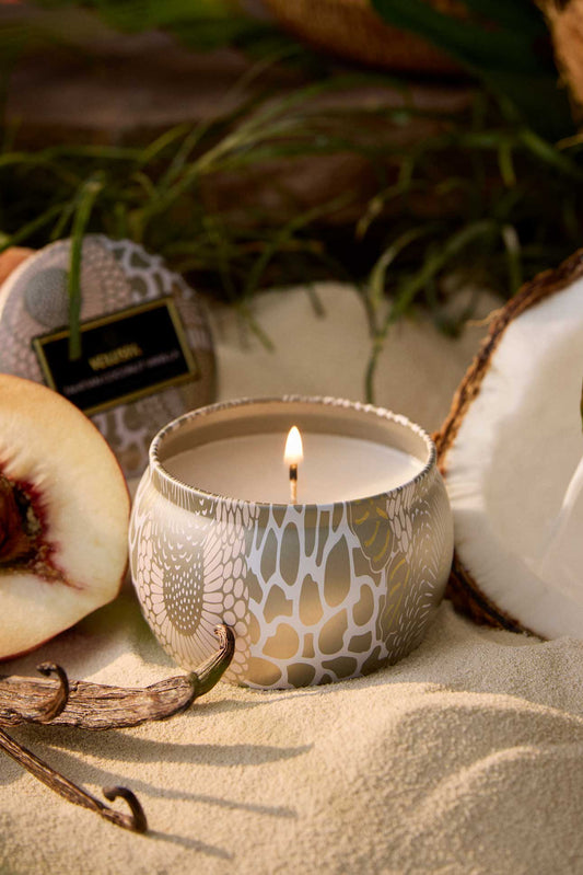 Candle in a decorative holder with peaches, a coconut, and a vanilla bean on a sandy surface. Voluspa Tahitian Coconut Vanilla 4 oz Mini Tin Candle