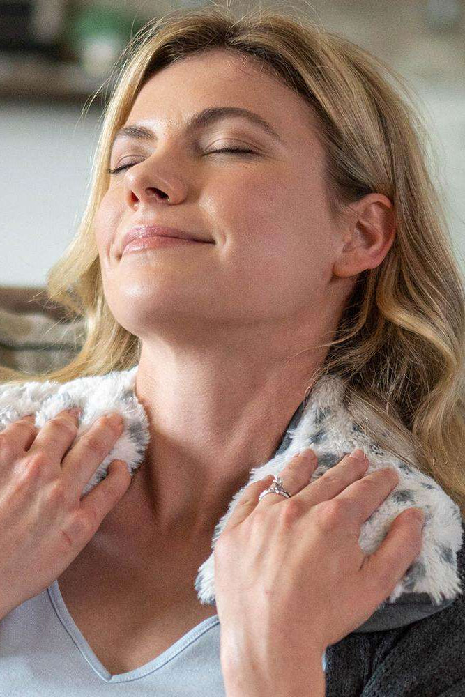 Woman wearing a gray robe with a fluffy collar, holding her neck and smiling.