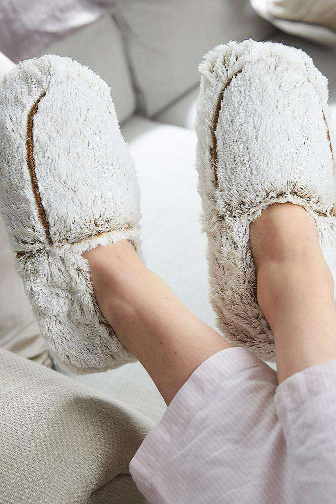 White fluffy slippers worn on a person's feet sitting on a light-colored couch.