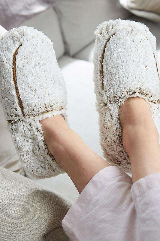 White fluffy slippers worn on a person's feet sitting on a light-colored couch.