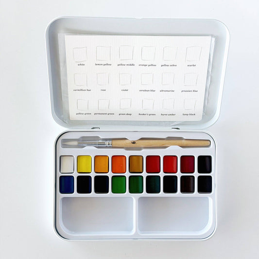 An open metal watercolor paint set with a white lid showing labeled color squares, a row of watercolor pans in assorted hues, a wooden-handled brush, and a small spatula inside a white case on a white background.