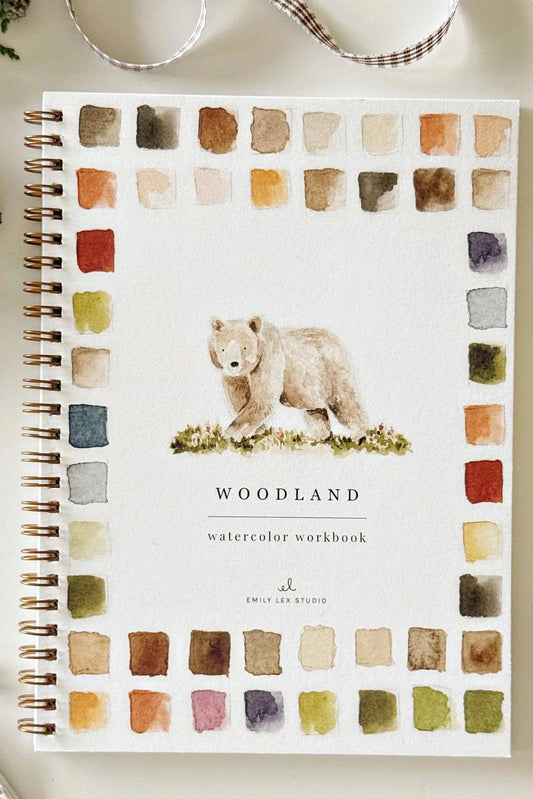 A spiral-bound watercolor workbook titled 'WOODLAND' with a bear illustration and watercolor swatches around the cover, styled on a light surface with a paintbrush, paint tube, and dried botanicals.