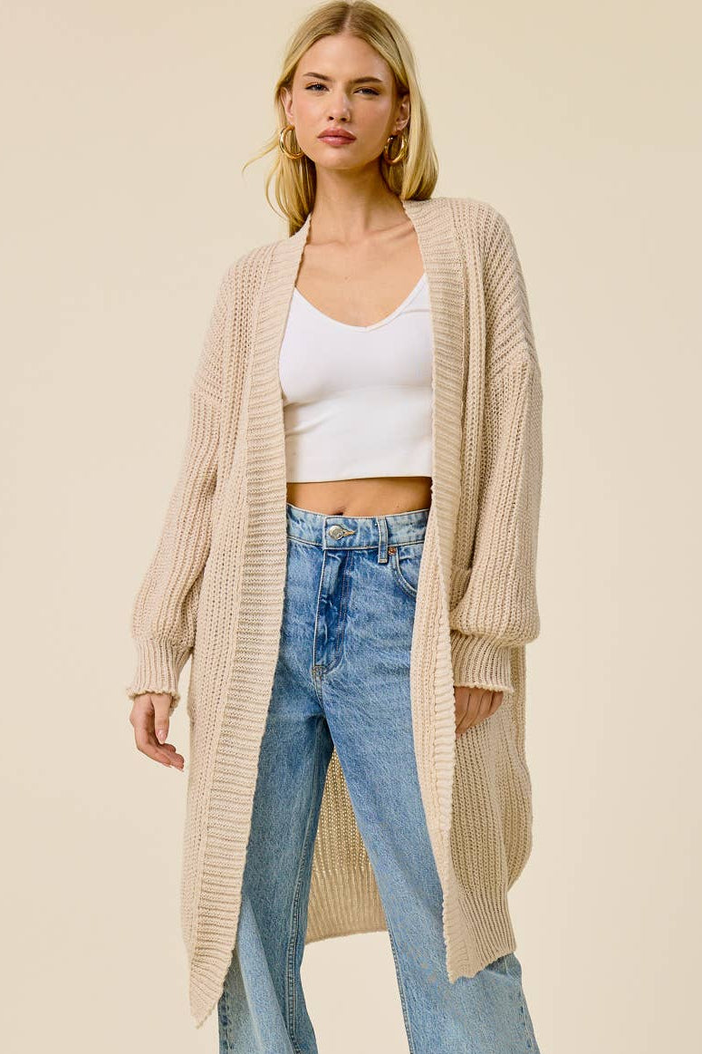 A woman modeling a long, open-front beige ribbed knit cardigan over a white crop top and light blue wide-leg jeans, photographed against a plain warm beige studio background.