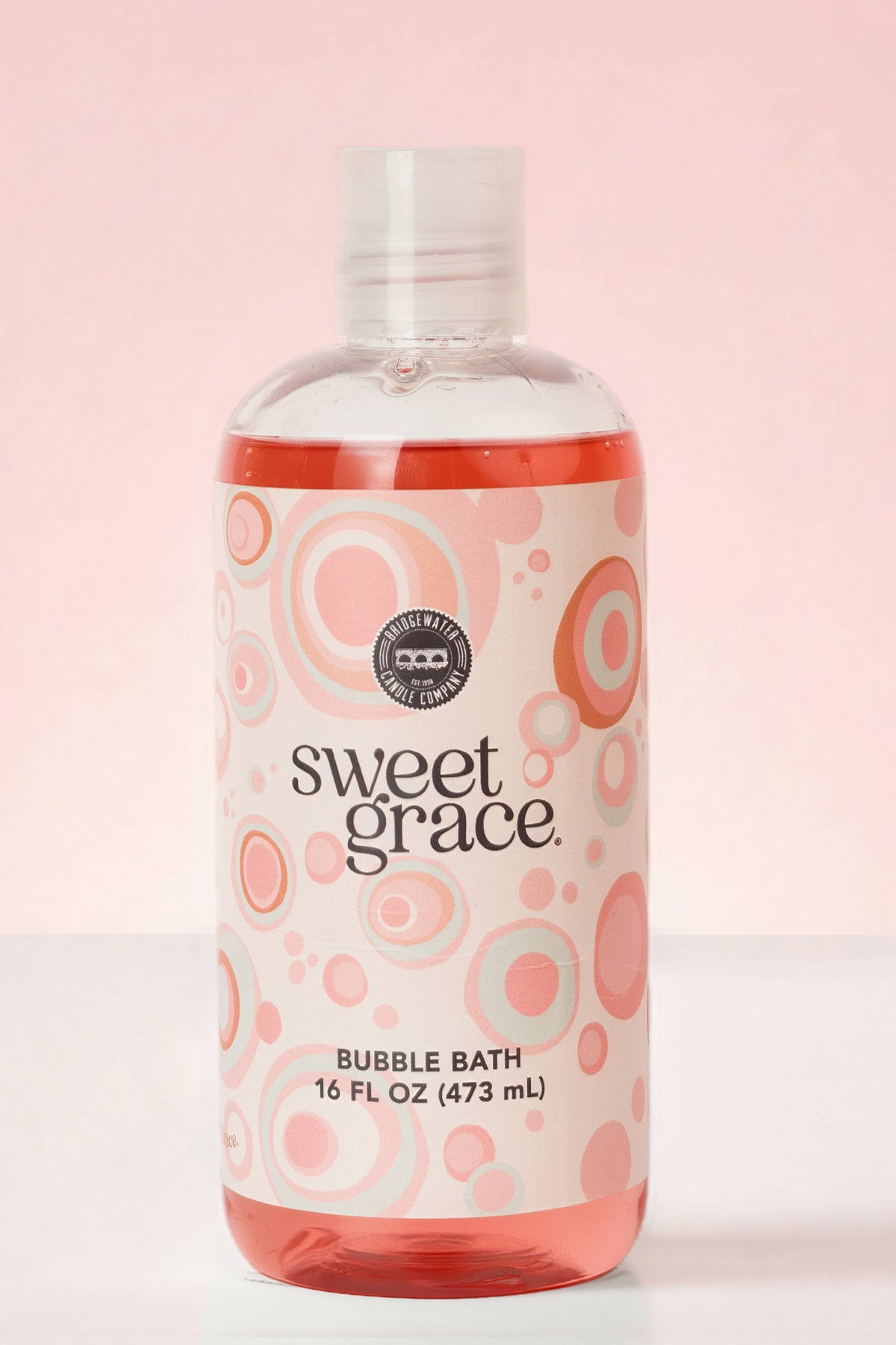 A 16 fl oz bottle of Sweet Grace Bubble Bath with pink and orange concentric circle patterns on the label, set against a soft pink background.