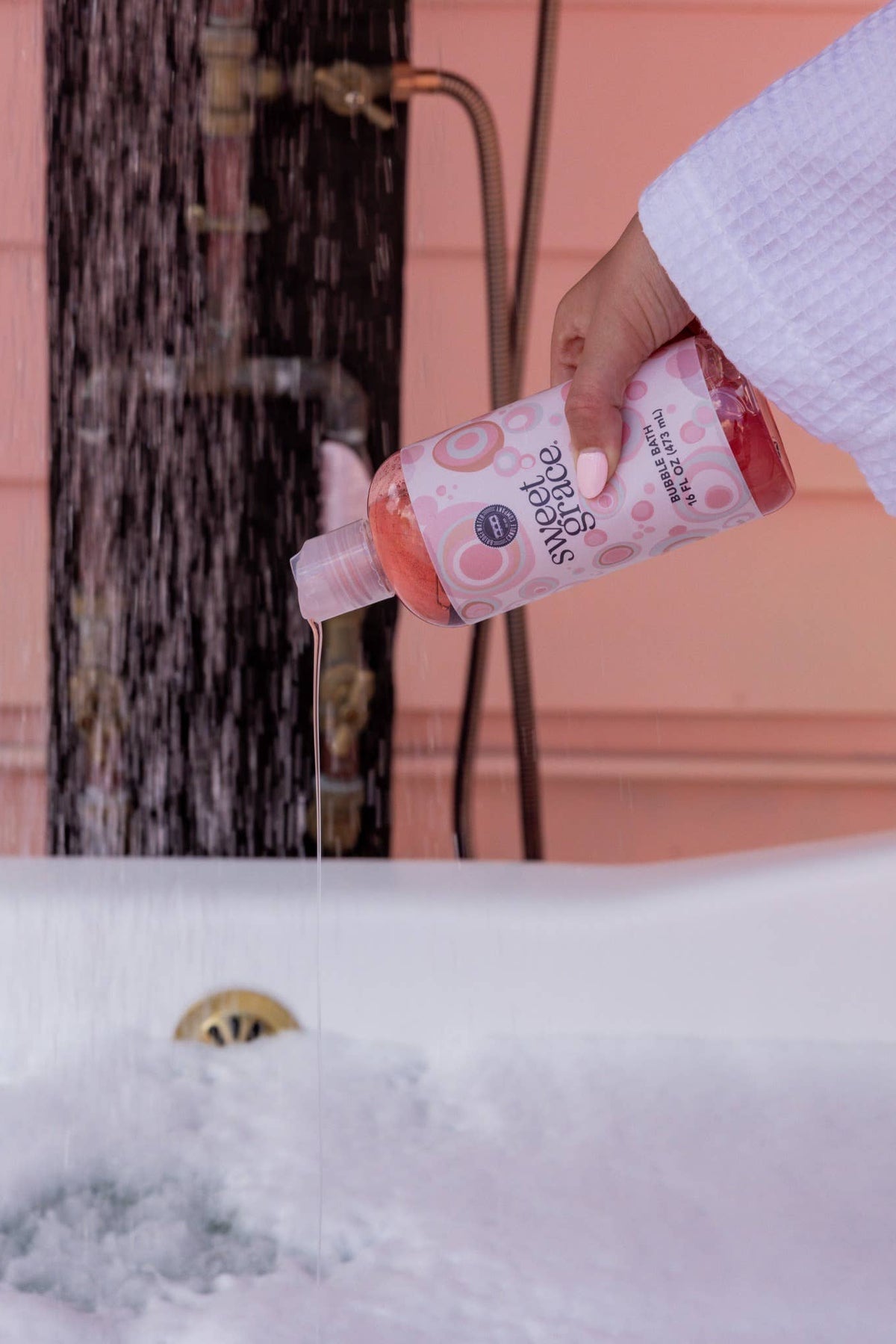 A hand in a white robe pours pink bath bomb powder from a bottle labeled 'Bath Time' into a bubbling bathtub with water running overhead.