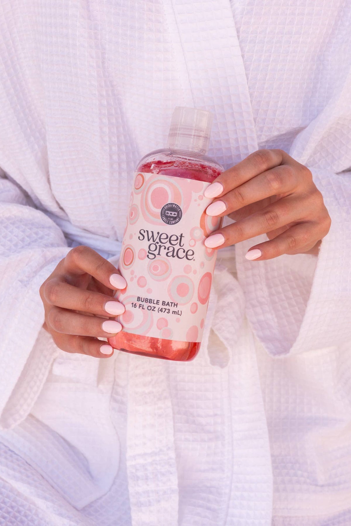 A person in a white waffle-textured robe holds a bottle of Sweet Grace Bubble Bath. The bottle is filled with pink liquid and has a pink label with white polka dots and the brand name 'sweet grace' in black script.