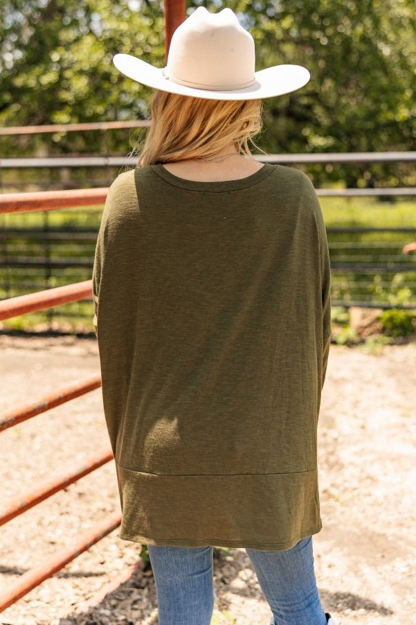 Person wearing a green sweater and white hat standing in an outdoor setting with trees and a fence.