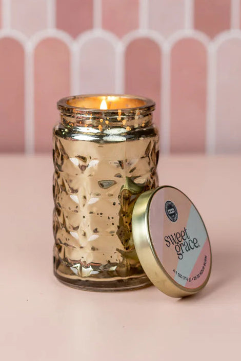 Gold candle jar with lid labeled 'Sweet Grace' on a pink background